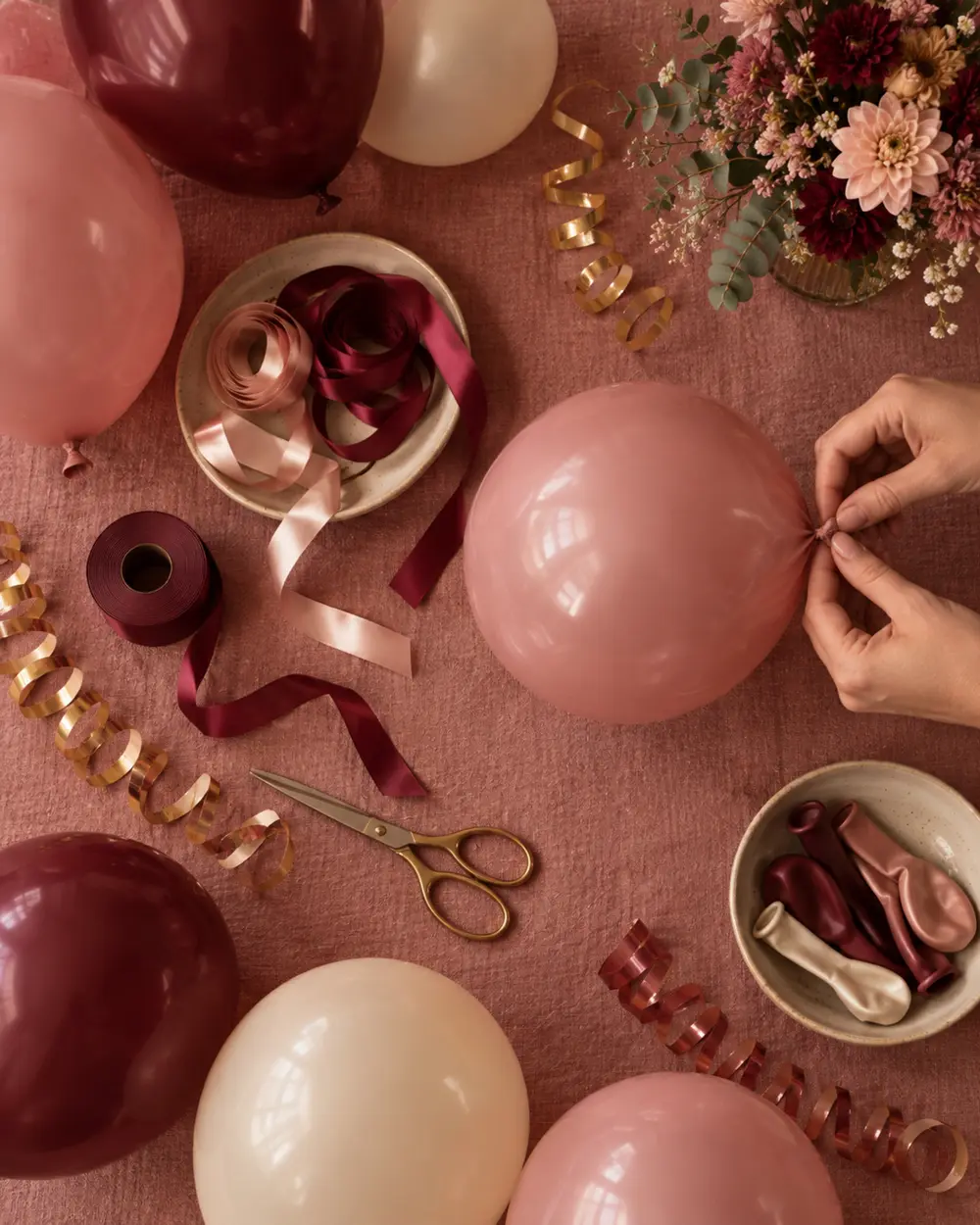 Mãos amarrando um balão rosa sobre a toalha, entre fitas, serpentina e balões vinho e creme.